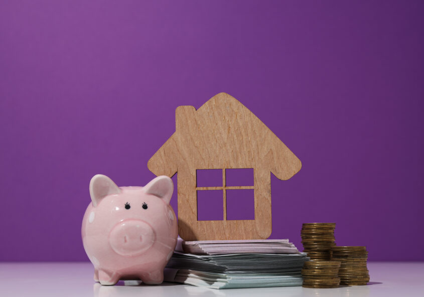 Wooden house, money, and piggy bank on purple background